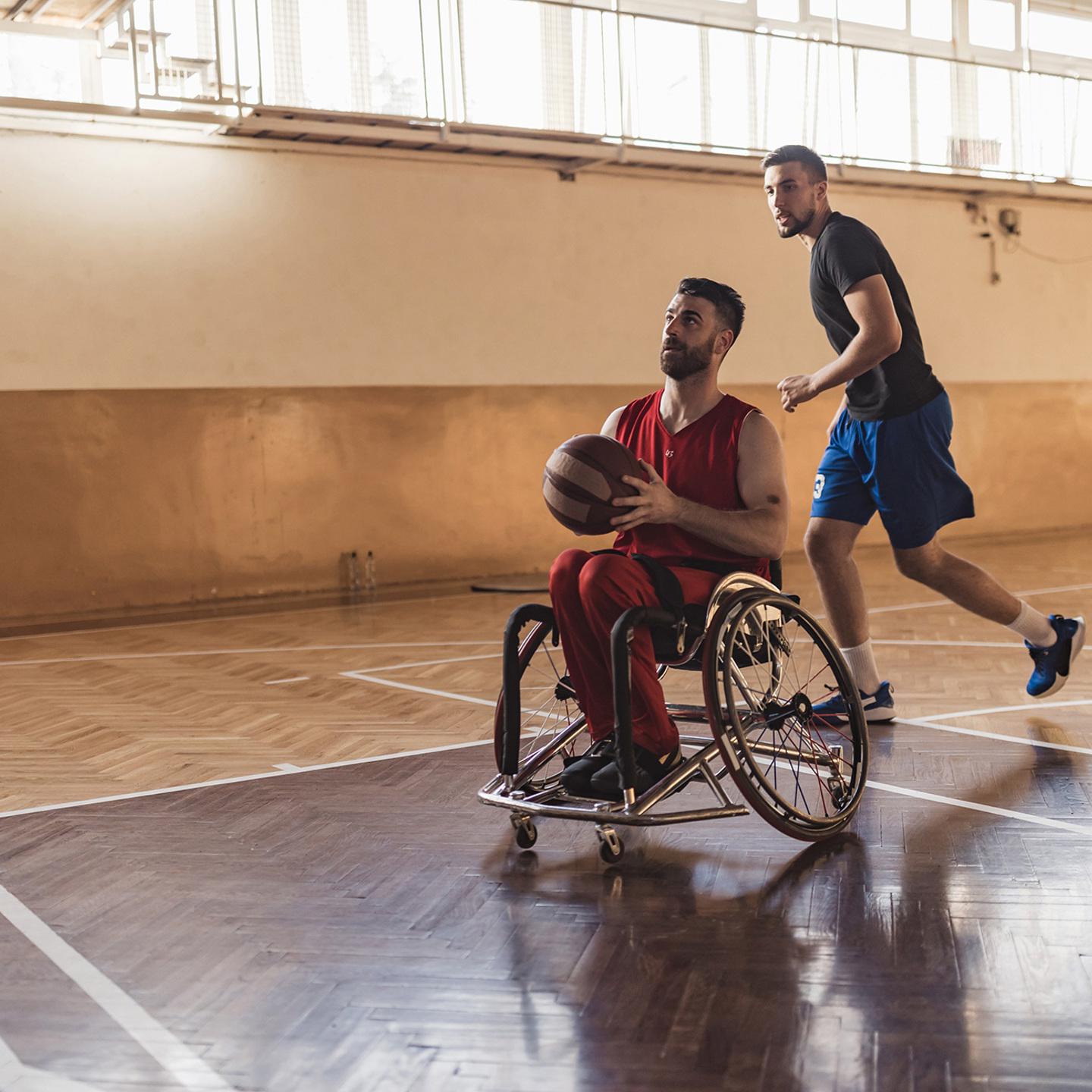 Photo of a person in a wheelchair playing basket ball in a sports hall with another person running behind them