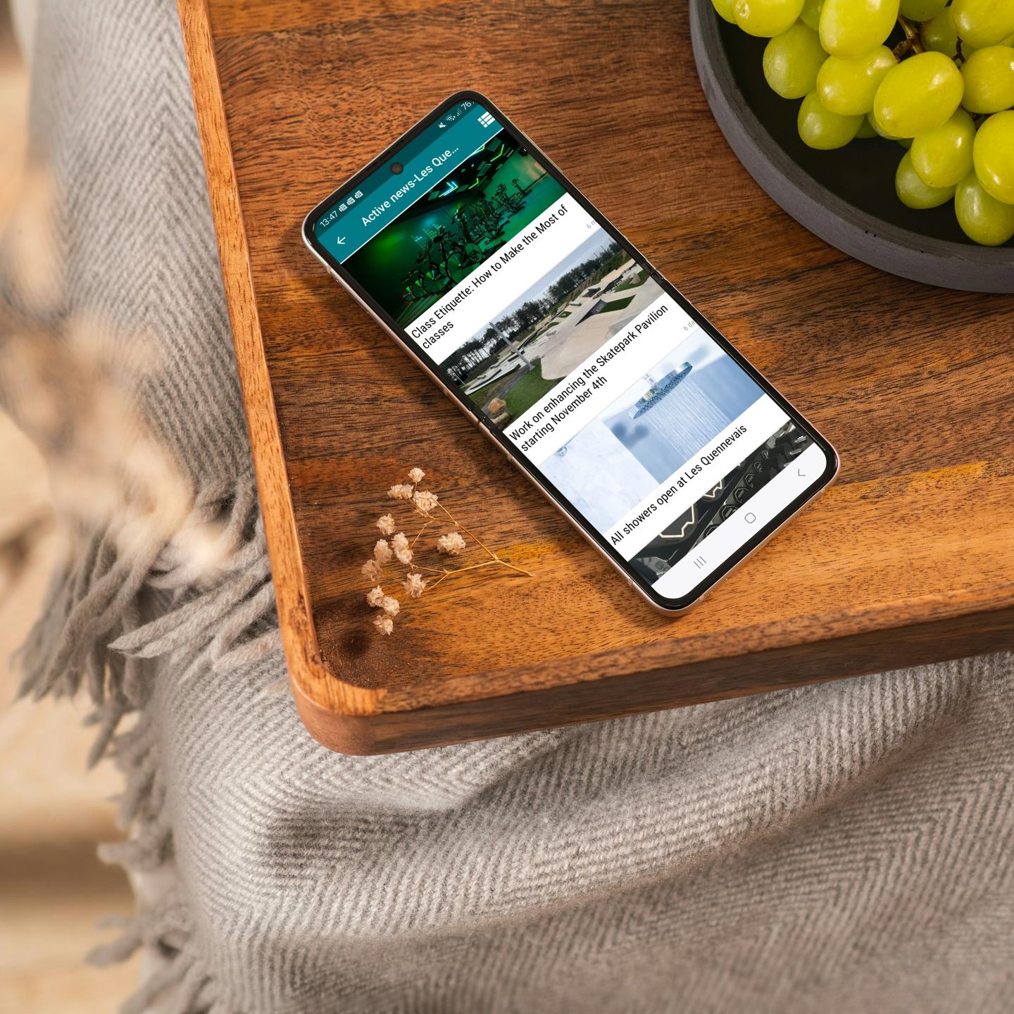 photo of the news section of the active app open on a mobile phone that is placed on a wooden table with a bowl of grapes next to it