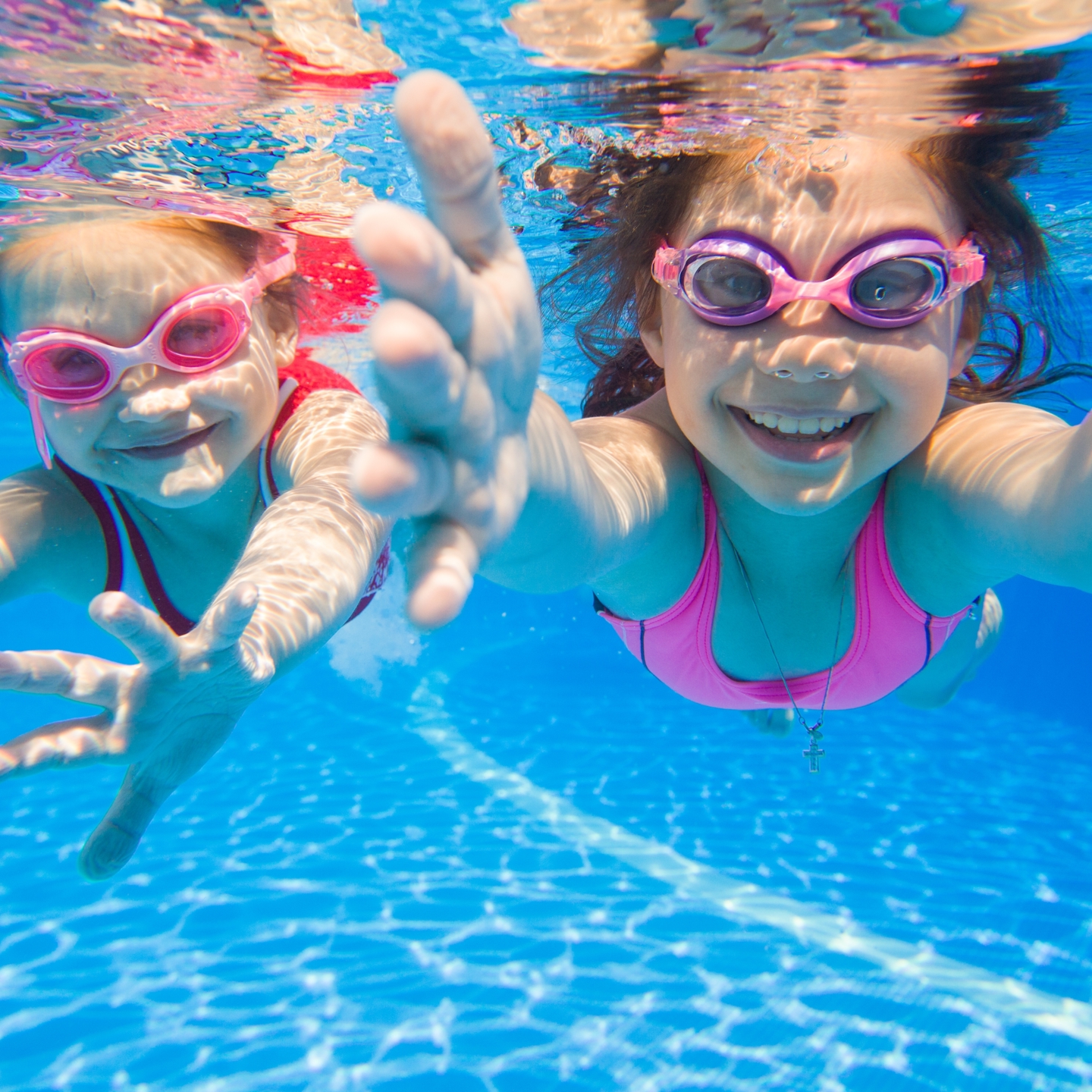 Photo of two children swimming underwater looking towards the camera with their arms outstretched reaching for it in a playful way