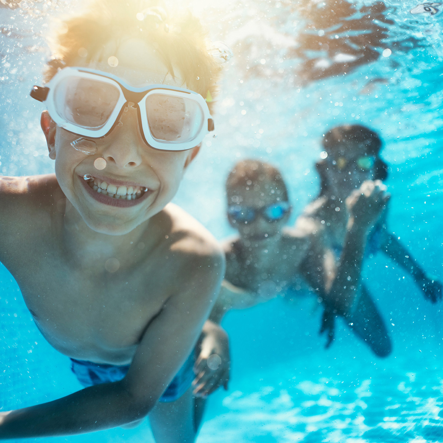 Smiling child with goggles on swimming underwater, two other children in bacground