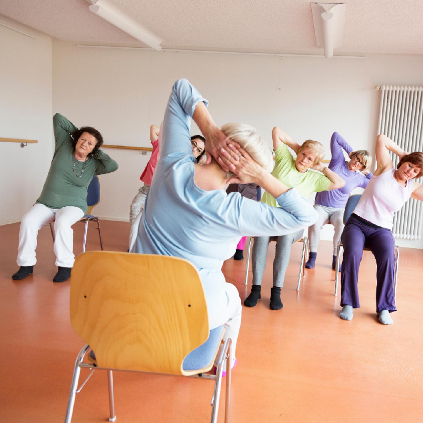 Photo of people taking part in a seated fitness class, the instructor has their back to the camera
