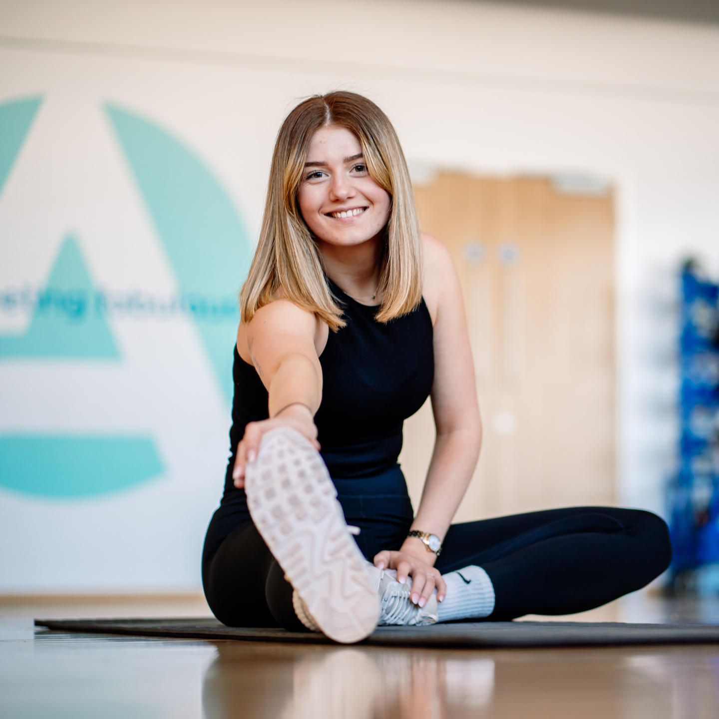 Photo of a woman in a studio on a matt stretching sat next to some weights and a kettle bell, you can see the active logo as well as some boxing gloves in he background